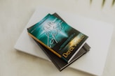 A book titled 'The Gifts of the Spirit' by Derek Prince is placed on top of a closed laptop and another notebook with an elastic band. The book cover features a serene, spiritual design with teal and golden hues, depicting a foggy landscape scene with a path leading toward a distant light.
