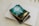 A book titled 'The Gifts of the Spirit' by Derek Prince is placed on top of a closed laptop and another notebook with an elastic band. The book cover features a serene, spiritual design with teal and golden hues, depicting a foggy landscape scene with a path leading toward a distant light.
