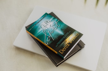A book titled 'The Gifts of the Spirit' by Derek Prince is placed on top of a closed laptop and another notebook with an elastic band. The book cover features a serene, spiritual design with teal and golden hues, depicting a foggy landscape scene with a path leading toward a distant light.