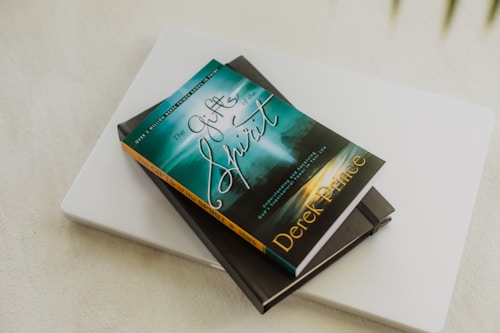 A book titled 'The Gifts of the Spirit' by Derek Prince is placed on top of a closed laptop and another notebook with an elastic band. The book cover features a serene, spiritual design with teal and golden hues, depicting a foggy landscape scene with a path leading toward a distant light.