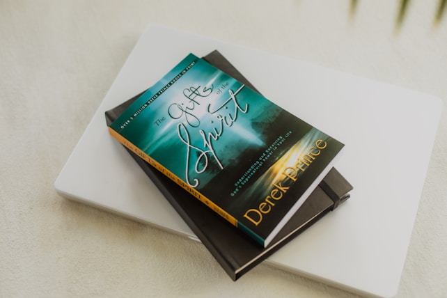 A book titled 'The Gifts of the Spirit' by Derek Prince is placed on top of a closed laptop and another notebook with an elastic band. The book cover features a serene, spiritual design with teal and golden hues, depicting a foggy landscape scene with a path leading toward a distant light.