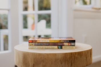 A wooden table holds a stack of three books by Derek Prince, arranged in a well-lit room with large windows in the background, allowing natural light to flood the space.