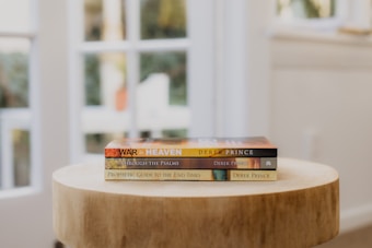 A wooden table holds a stack of three books by Derek Prince, arranged in a well-lit room with large windows in the background, allowing natural light to flood the space.