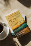 A book titled 'Prophetic Guide to the End Times' by Derek Prince lies on a marble surface. Next to the book, there is a white cup filled with coffee placed on a saucer. Sunlight casts soft shadows on the surface.