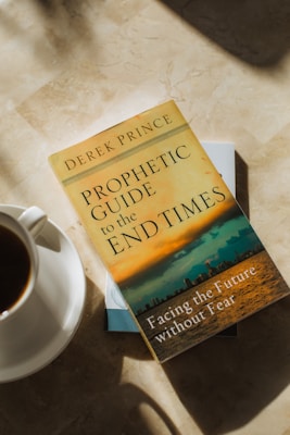 A book titled 'Prophetic Guide to the End Times' by Derek Prince lies on a marble surface. Next to the book, there is a white cup filled with coffee placed on a saucer. Sunlight casts soft shadows on the surface.