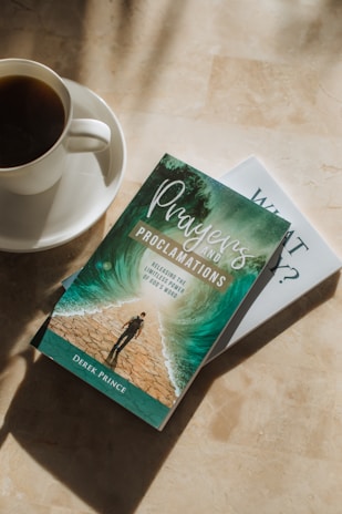 A cup of coffee sits on a saucer next to two books placed on a sunlit surface. The top book has a cover depicting a person walking between parted waters, with the title 'Prayers and Proclamations' by Derek Prince. The lighting casts soft shadows on the surface.