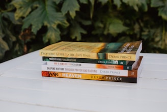 A stack of five books rests on a white wooden surface. Each book cover features text and distinct designs, with titles like 'Prophetic Guide to the End Times' and 'War in Heaven' by Derek Prince. The background consists of lush green leaves, offering a natural backdrop to the scene.