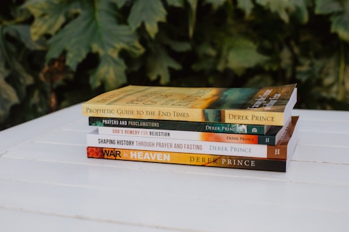 A stack of five books rests on a white wooden surface. Each book cover features text and distinct designs, with titles like 'Prophetic Guide to the End Times' and 'War in Heaven' by Derek Prince. The background consists of lush green leaves, offering a natural backdrop to the scene.