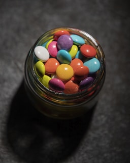 A glass jar filled with an assortment of colorful candy-coated chocolates sits on a textured dark surface. The candies are round and come in various vibrant colors including red, blue, pink, yellow, green, purple, and white. The lighting casts a soft shadow around the jar, enhancing the vividness of the candies.