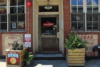 The image shows the facade of a restaurant with brick walls and a wooden door. There are signs advertising seafood like crab and shrimp. A neon sign in the window reads 'Extra Fancy,' and there are planters with greenery on either side of the entrance. A notice indicates that the kitchen is open until 2.