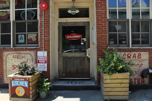 The image shows the facade of a restaurant with brick walls and a wooden door. There are signs advertising seafood like crab and shrimp. A neon sign in the window reads 'Extra Fancy,' and there are planters with greenery on either side of the entrance. A notice indicates that the kitchen is open until 2.