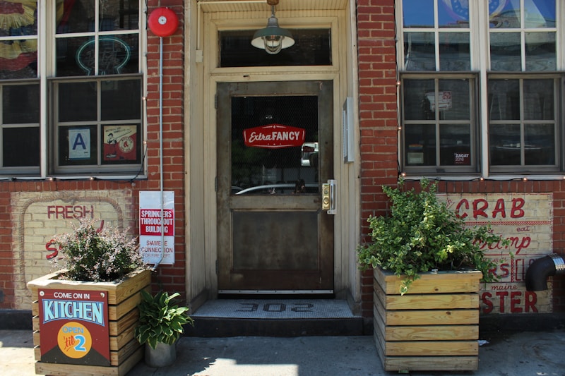 The image shows the facade of a restaurant with brick walls and a wooden door. There are signs advertising seafood like crab and shrimp. A neon sign in the window reads 'Extra Fancy,' and there are planters with greenery on either side of the entrance. A notice indicates that the kitchen is open until 2.