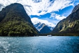 A scenic view of a fjord with towering green mountains on either side, a calm body of water in the foreground, and a small boat floating in the center. Snow-capped peaks are visible in the distance under a partly cloudy sky.
