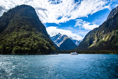 A scenic view of a fjord with towering green mountains on either side, a calm body of water in the foreground, and a small boat floating in the center. Snow-capped peaks are visible in the distance under a partly cloudy sky.