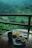 A serene view of lush green hills and rice paddies seen from a balcony, with a wooden table in the foreground featuring a mug, a plate of pancakes, a bowl of sauce, and a dish of fries.