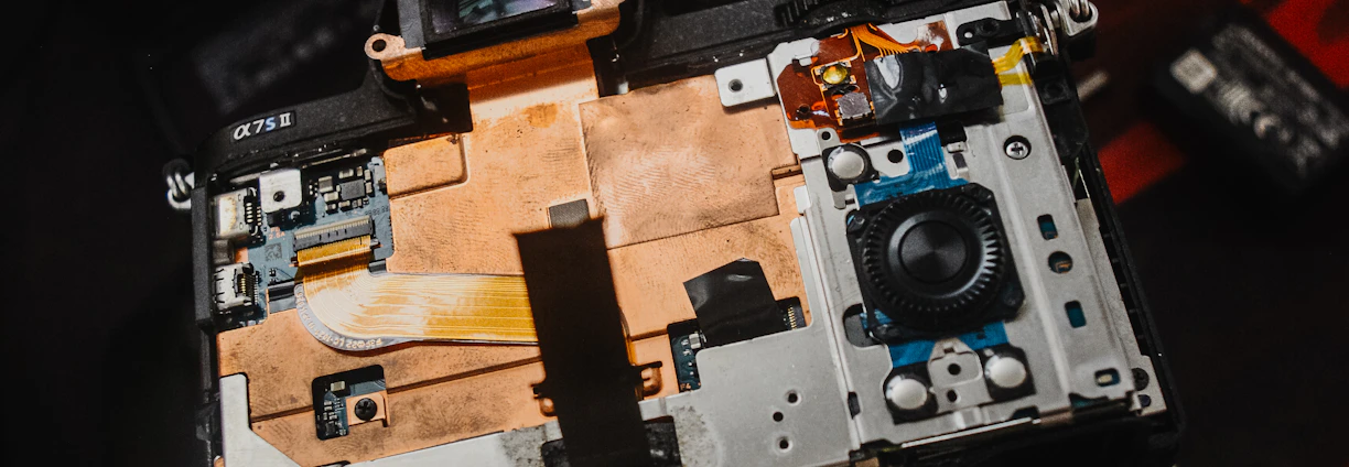 An open electronic device with visible internal components, including circuitry and connectors. The device rests on a dark surface with a logo and another small gadget visible nearby, indicating a focus on technology and repair.