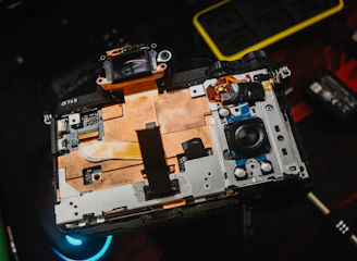 An open electronic device with visible internal components, including circuitry and connectors. The device rests on a dark surface with a logo and another small gadget visible nearby, indicating a focus on technology and repair.