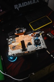 An open electronic device with visible internal components, including circuitry and connectors. The device rests on a dark surface with a logo and another small gadget visible nearby, indicating a focus on technology and repair.