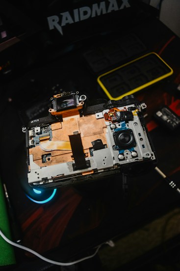 An open electronic device with visible internal components, including circuitry and connectors. The device rests on a dark surface with a logo and another small gadget visible nearby, indicating a focus on technology and repair.