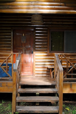 A wooden cabin with a front porch featuring a set of stairs leading up to a door marked with the number 1. The cabin has a log exterior, a lantern hanging above the door, a window with the curtain partly drawn, and a blue bin to the left of the stairs.