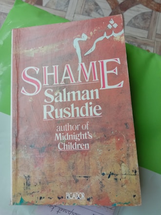 A book titled 'Shame' authored by Salman Rushdie is placed on a bright green surface. The book cover features a mix of earthy tones with abstract textures and large white typography. Arabic script is displayed at the top part of the cover.