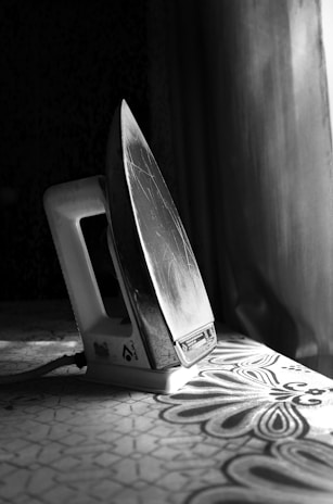 A vintage-style clothes iron is positioned upright on a textured surface, with light streaming in from a nearby window. The iron's metal soleplate shows visible scratches, indicating frequent use. The base of the iron bears a brand logo and some text, while the background is a blend of darkness and soft light.