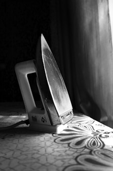 A vintage-style clothes iron is positioned upright on a textured surface, with light streaming in from a nearby window. The iron's metal soleplate shows visible scratches, indicating frequent use. The base of the iron bears a brand logo and some text, while the background is a blend of darkness and soft light.