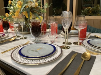 A beautifully set dining table with elegant tableware, including decorative plates, gold utensils, and an assortment of glassware for various beverages. The centerpiece is a floral arrangement with yellow and white flowers, adding a touch of elegance to the setting. Drinks of different colors, including red and dark, fill the glasses, enhancing the luxurious feel.
