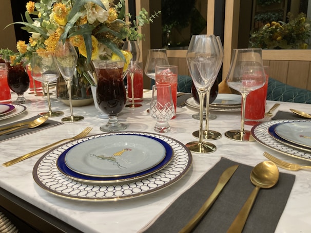 A beautifully set dining table with elegant tableware, including decorative plates, gold utensils, and an assortment of glassware for various beverages. The centerpiece is a floral arrangement with yellow and white flowers, adding a touch of elegance to the setting. Drinks of different colors, including red and dark, fill the glasses, enhancing the luxurious feel.