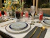 A beautifully set dining table with elegant tableware, including decorative plates, gold utensils, and an assortment of glassware for various beverages. The centerpiece is a floral arrangement with yellow and white flowers, adding a touch of elegance to the setting. Drinks of different colors, including red and dark, fill the glasses, enhancing the luxurious feel.