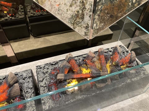 Logs arranged in a decorative fireplace setup. The logs have red and orange accents, simulating glowing embers, and are surrounded by a metal or stone frame. The setup is behind a glass panel, reflecting light and creating a warm ambiance.
