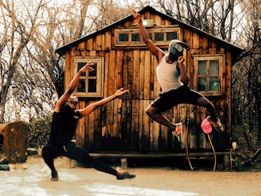 Two people are engaged in a dynamic and expressive dance performance in front of a rustic wooden cabin. One dancer is in a low, wide stance with arms outstretched towards the other dancer, who is captured in mid-air, wearing a helmet and making an energetic gesture with one arm. The background is filled with bare trees, giving a rustic and natural ambiance.
