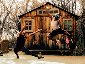 Two people are engaged in a dynamic and expressive dance performance in front of a rustic wooden cabin. One dancer is in a low, wide stance with arms outstretched towards the other dancer, who is captured in mid-air, wearing a helmet and making an energetic gesture with one arm. The background is filled with bare trees, giving a rustic and natural ambiance.