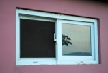 A rectangular window with white framing set in a pink wall. On the right pane, there is a reflection of a scenic view with tree silhouettes and a sunset sky. The left pane appears dark, possibly a screen or tinted glass.