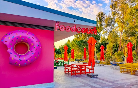 A vibrant outdoor food court area featuring a large pink wall with a giant donut decoration. The seating area includes brightly colored chairs and tables, mainly in red and yellow, with orange umbrellas providing shade. The setting is surrounded by lush greenery and trees under a partly cloudy sky.
