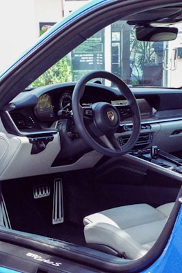 The interior of a luxury car featuring a sleek steering wheel with a prominent logo, a modern dashboard, and a comfortable light-colored seat. The car door is open, revealing a clear view of the interior design.