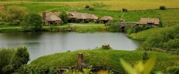 A serene landscape featuring a small lake surrounded by lush greenery. Quaint thatched-roof cottages are nestled on the far side of the water, accompanied by a small stone bridge. The rolling hills in the background enhance the pastoral setting, and a well-kept lawn extends to the foreground.