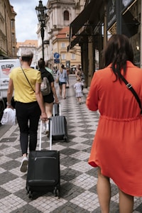 People walking down a cobblestone street in a historic city, with some pulling suitcases. The street is lined with buildings featuring classic architecture, and there are signs of commerce and a van parked nearby.