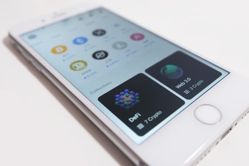 A smartphone displays a cryptocurrency app interface with logos of different cryptocurrencies like Bitcoin, Dogecoin, and Stellar. The app includes sections for various collections like DeFi and Web 3.0, with the number of cryptos listed under each.