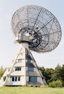 A large radio telescope with a wire-mesh dish and a geometric base structure, set against a clear blue sky. The base resembles a building with windows, surrounded by greenery and a grass field.