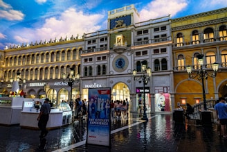 A large, ornate shopping center with architecture resembling Venetian buildings, featuring multiple levels with arched windows and classic street lamps. The ground is wet as though from rain, reflecting the warm lighting from the buildings. There is a clock tower in the center, with brands like Pandora and Alex and Ani displayed prominently. Shoppers are walking around, adding a lively atmosphere.