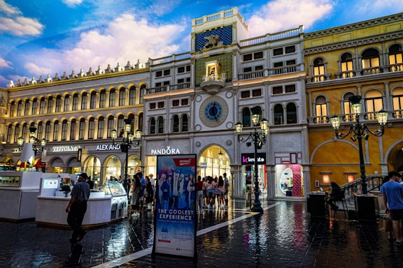 A large, ornate shopping center with architecture resembling Venetian buildings, featuring multiple levels with arched windows and classic street lamps. The ground is wet as though from rain, reflecting the warm lighting from the buildings. There is a clock tower in the center, with brands like Pandora and Alex and Ani displayed prominently. Shoppers are walking around, adding a lively atmosphere.