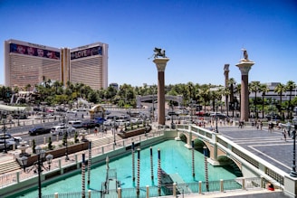 A view of a bustling urban area with a prominent hotel featuring large advertisements. The scene includes a wide canal with gondola poles, surrounded by classical-style columns topped with statues. There is a mix of cars and pedestrians on the streets, and abundant greenery including palm trees.