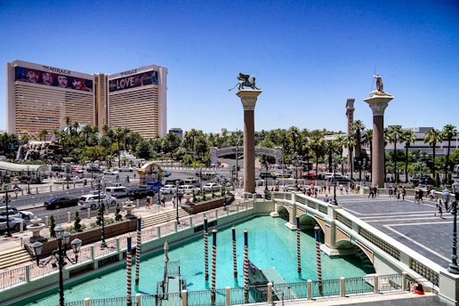 A view of a bustling urban area with a prominent hotel featuring large advertisements. The scene includes a wide canal with gondola poles, surrounded by classical-style columns topped with statues. There is a mix of cars and pedestrians on the streets, and abundant greenery including palm trees.
