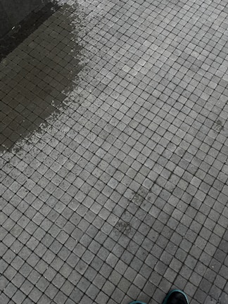 A tiled surface composed of small, square tiles in a grid pattern. There is a slight color variation indicating dampness on part of the surface. The corner of a shoe is visible at the bottom right corner.