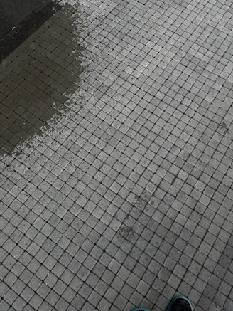 A tiled surface composed of small, square tiles in a grid pattern. There is a slight color variation indicating dampness on part of the surface. The corner of a shoe is visible at the bottom right corner.