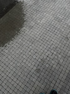 A tiled surface composed of small, square tiles in a grid pattern. There is a slight color variation indicating dampness on part of the surface. The corner of a shoe is visible at the bottom right corner.