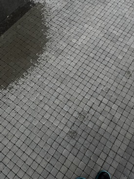 A tiled surface composed of small, square tiles in a grid pattern. There is a slight color variation indicating dampness on part of the surface. The corner of a shoe is visible at the bottom right corner.