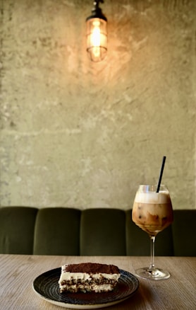 A slice of tiramisu sits on a dark plate on a wooden table, accompanied by a tall glass of iced coffee topped with foam. Above, a vintage-style bulb in a caged light fixture casts a warm glow against a textured stone wall. The setting conveys a cozy and inviting caf&eacute; atmosphere.