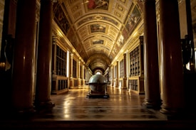 A grand, ornately decorated hallway lined with tall pillars and arched ceilings, featuring intricate artwork on the ceiling and walls. At the center, there is a large globe placed on a pedestal, surrounded by shelves filled with books, creating a sense of historical grandeur.
